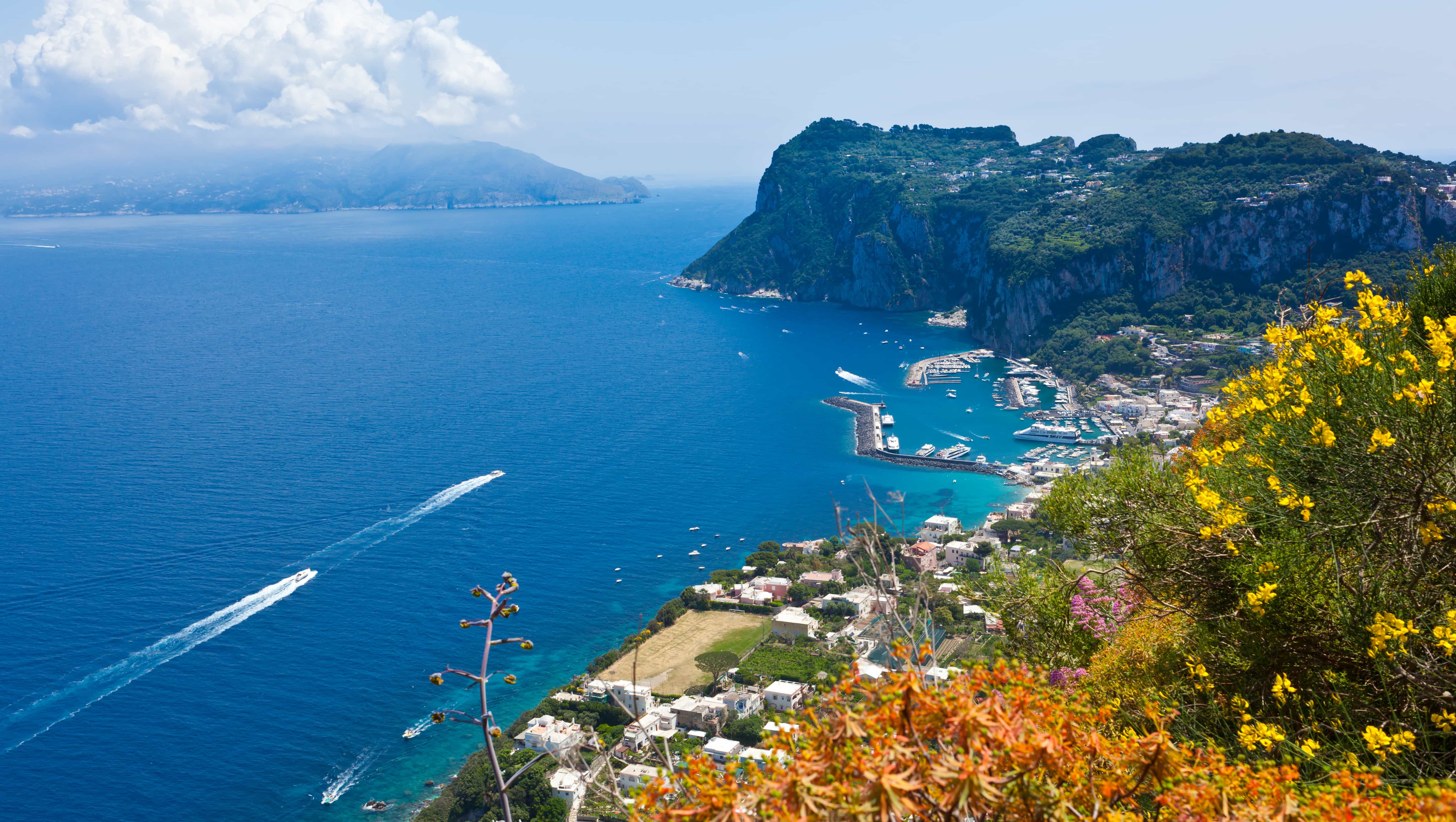 Bird's eye view of yachts sailing just off Italy's coast heading toward a small marina at the foot of a mountain