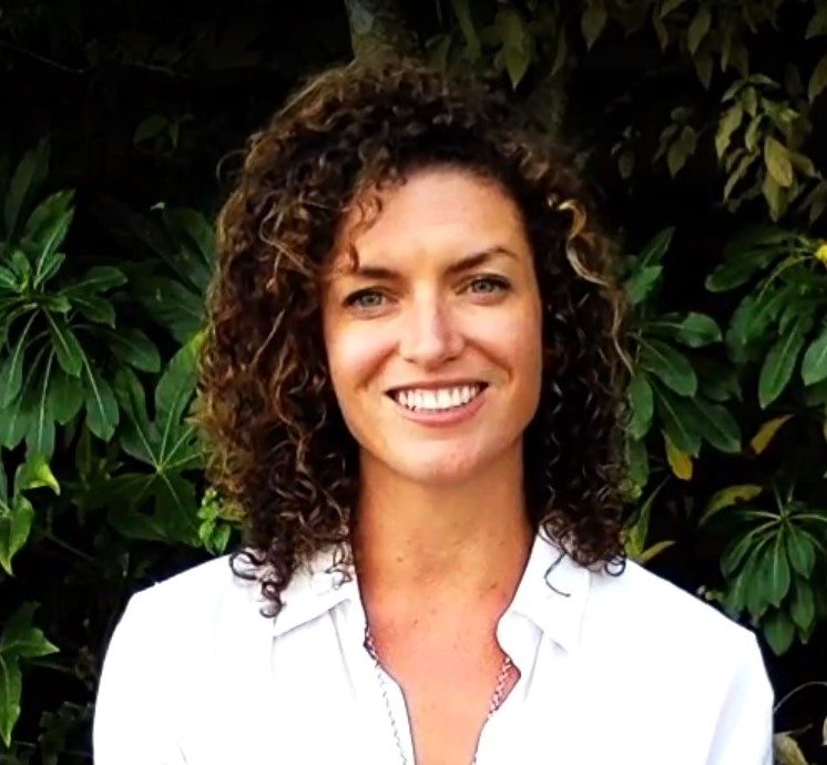 brunette with curly hair smiling in front of green bushes