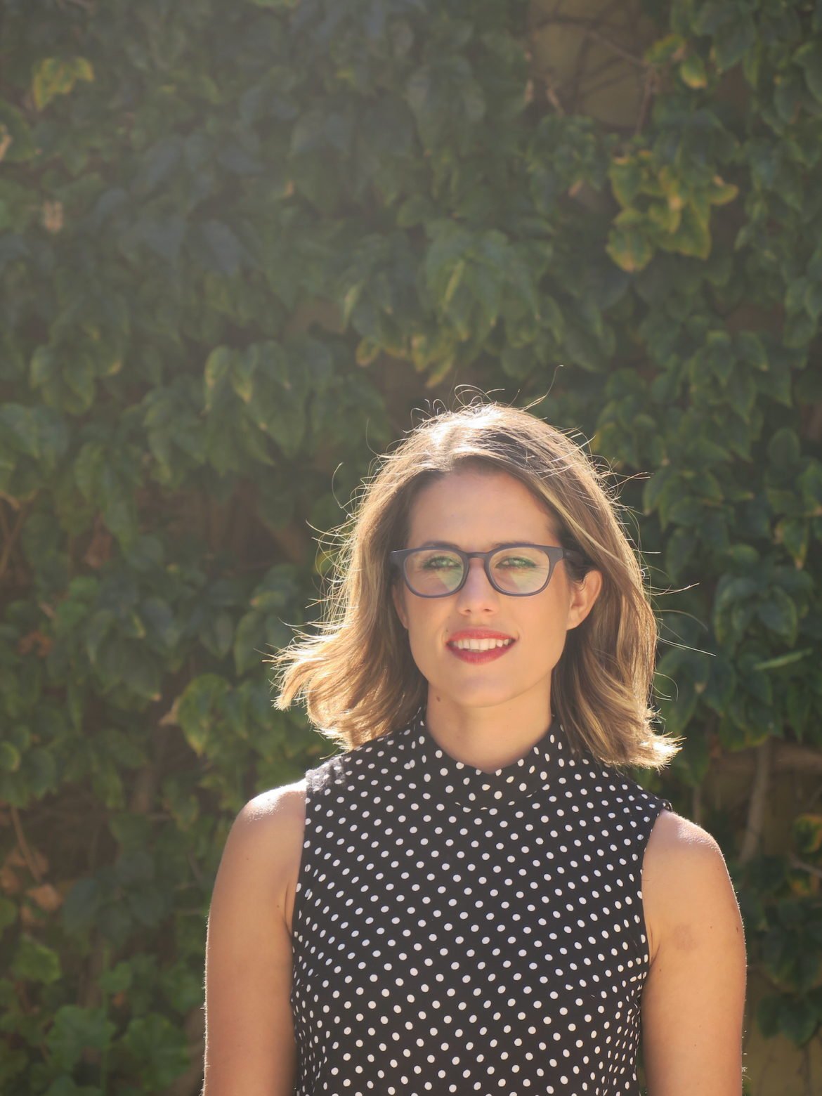 Karine Rayson smiling with short blonde hair, black glasses in front of large bush