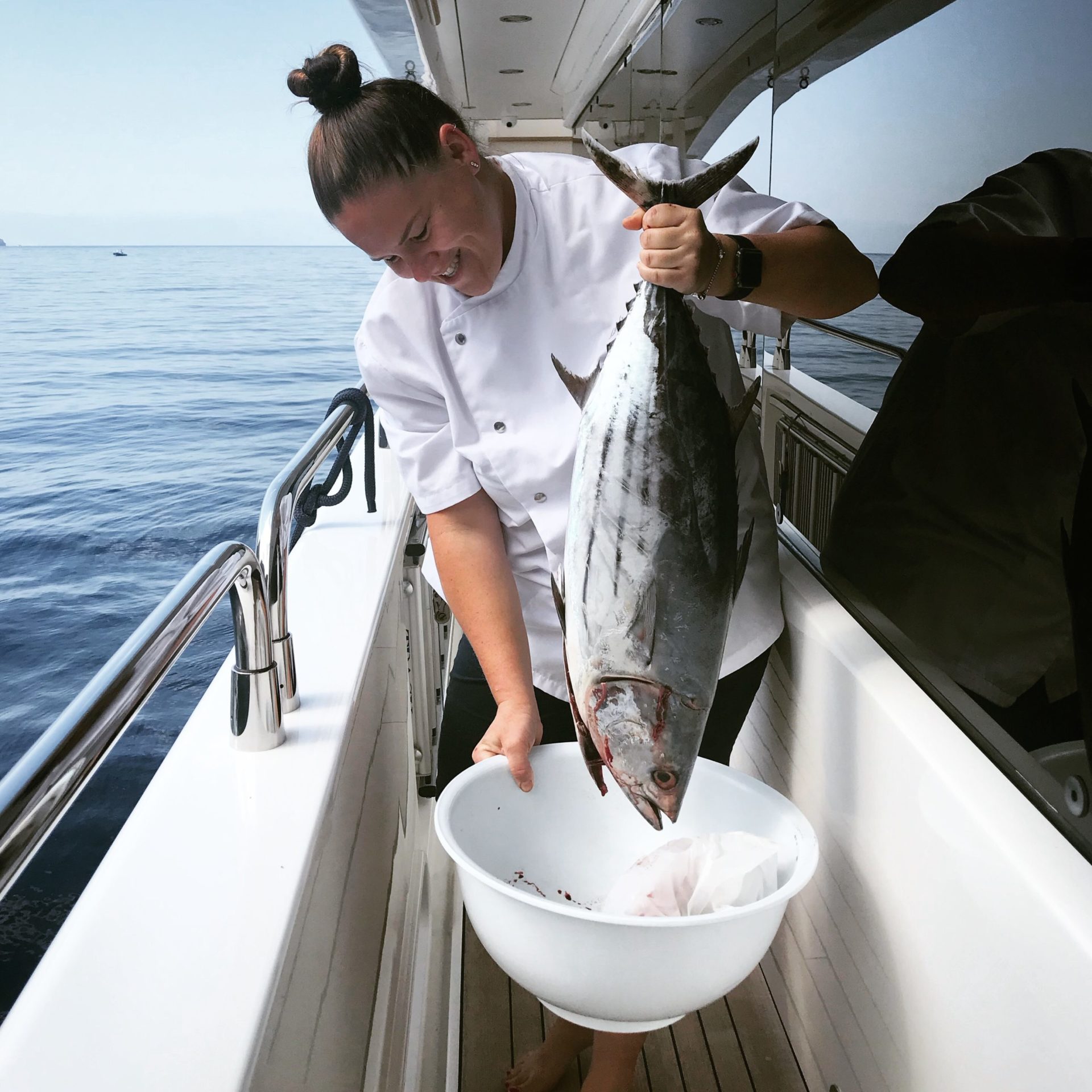 Chef Jeanri in chef jacket smiling down while holding large fish onboard a yacht