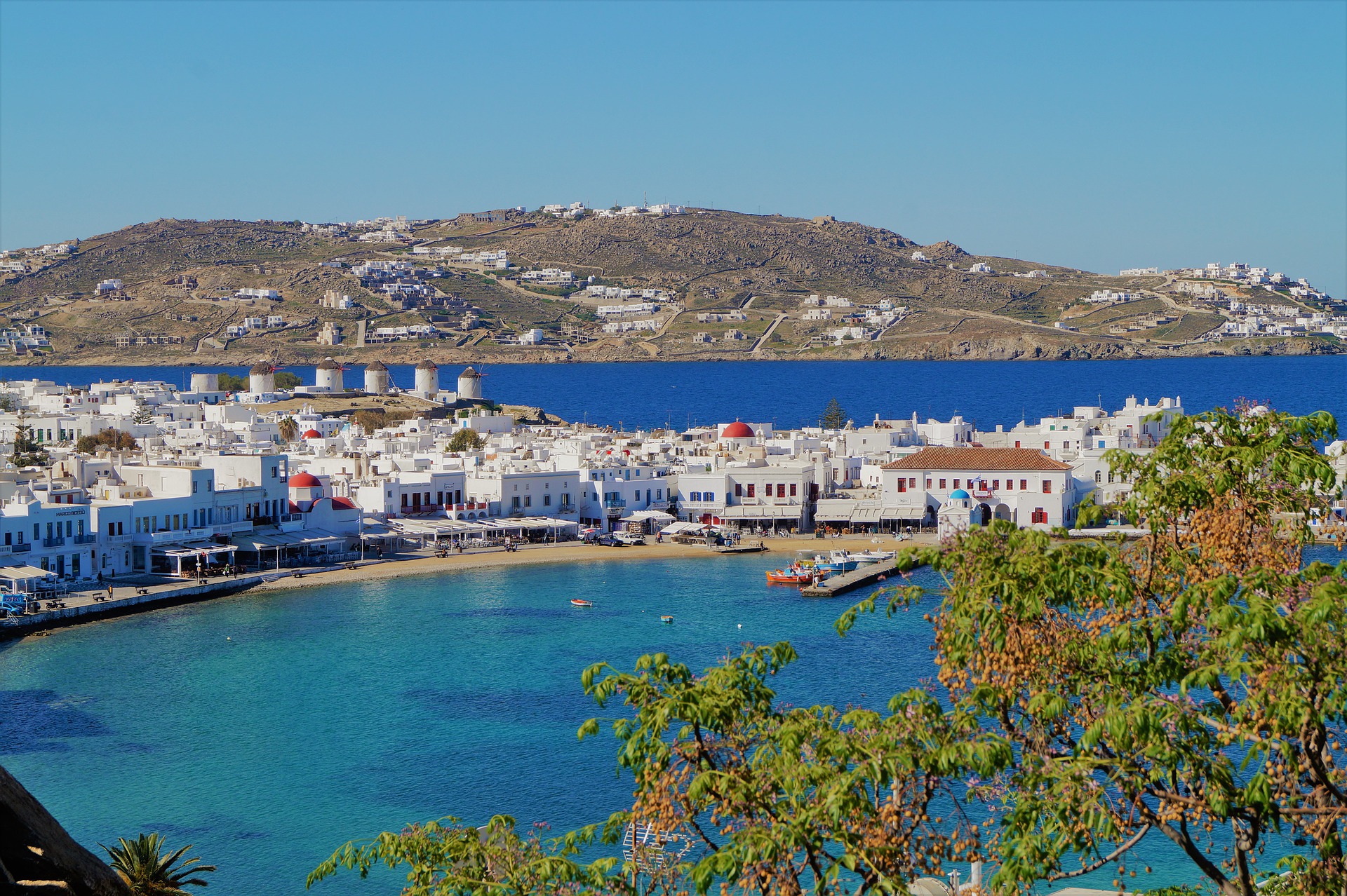 shot of Mykonos harbour with turquoise blue waters and white washed buildings with mountains in the background